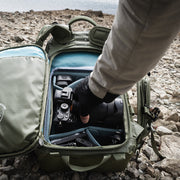 An image of the Shimoda Explore V2 25 Backpack Starter Kit in green. A photographer is pictured taking a camera out of the bag, which contains other photography equipment, against a rocky landscape.