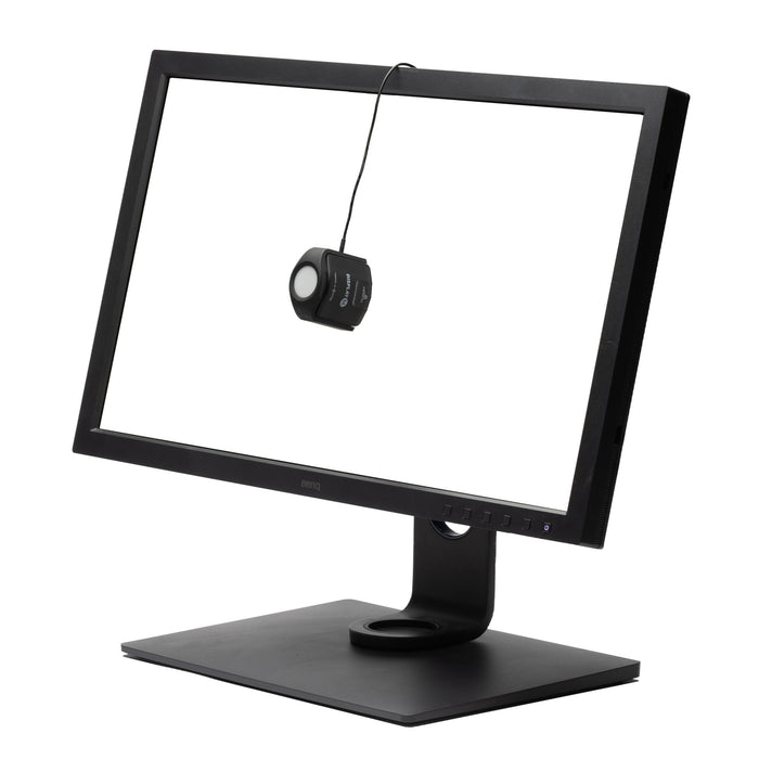 An image of the Calibrite Display SL calibration device, powered by X-Rite. It is hanging over a monitor screen by the cable so that the device can measure and calibrate the colours on screen.