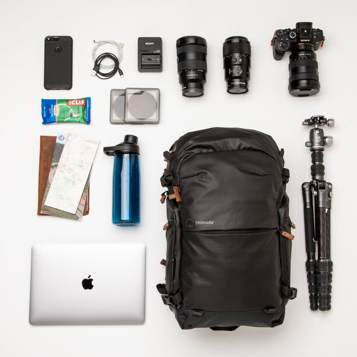 An image of the Shimoda Explore V2 25 Backpack Starter Kit Black. The backpack is black with rust coloured zips and has a carry handle, pockets for water bottles or tripods and multiple zipped pockets with shoulder straps and a removeable belt. The backpack is pictured next to a laptop, tripod, travel documents, water bottle, phone, cables, charger, lens filters, snacks, a camera and two lenses.