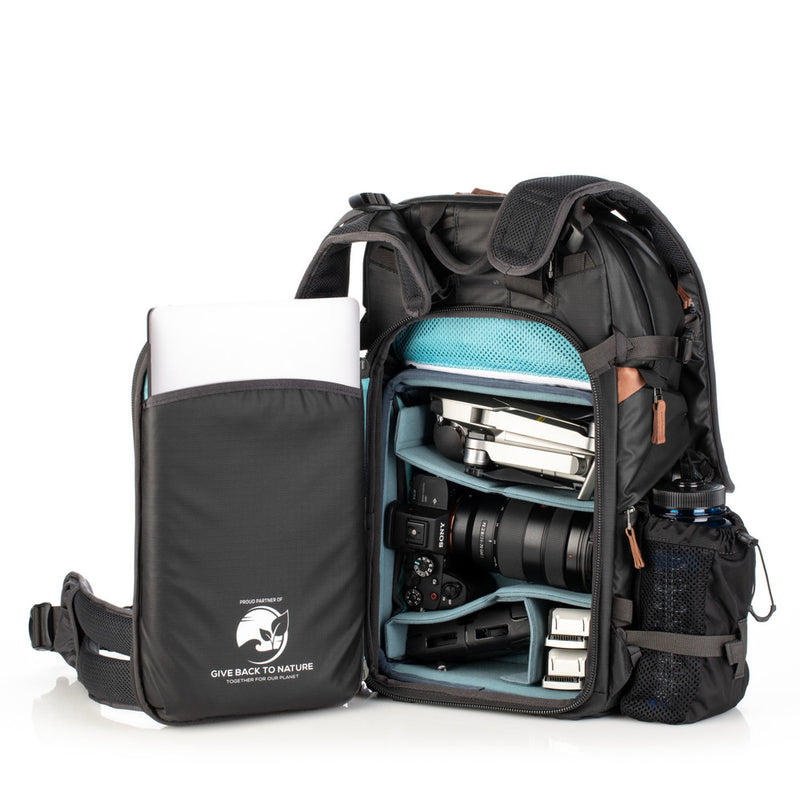 An image of the Shimoda Explore V2 25 Backpack Starter Kit Black with the back compartment open. The backpack is black with rust coloured zips and has a carry handle, pockets for water bottles or tripods and multiple zipped pockets with shoulder straps and a removeable belt. Inside the backpack is a laptop, water bottle, camera and other photographic equipment.