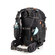 An image of the Shimoda Explore V2 25 Backpack Starter Kit Black with the shoulder strap compartment open. The backpack is black with rust coloured zips and has a carry handle, pockets for water bottles or tripods and multiple zipped pockets with shoulder straps and a removeable belt. Inside the shoulder strap is a piece of RODE photography equipment and there is a detachable belt for additional support.
