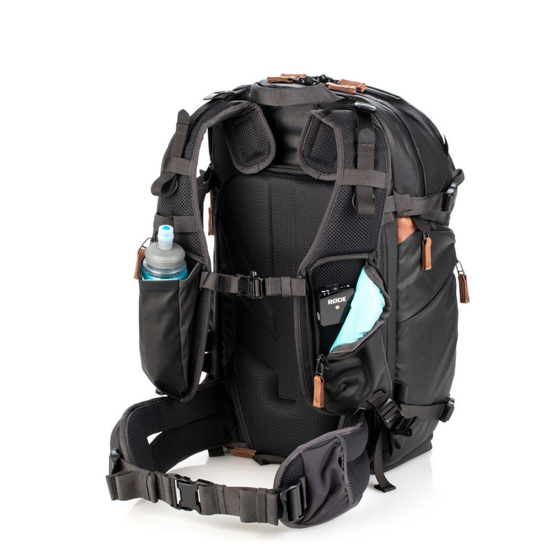 An image of the Shimoda Explore V2 25 Backpack Starter Kit Black with the shoulder strap compartment open. The backpack is black with rust coloured zips and has a carry handle, pockets for water bottles or tripods and multiple zipped pockets with shoulder straps and a removeable belt. Inside the shoulder strap is a piece of RODE photography equipment and there is a detachable belt for additional support.